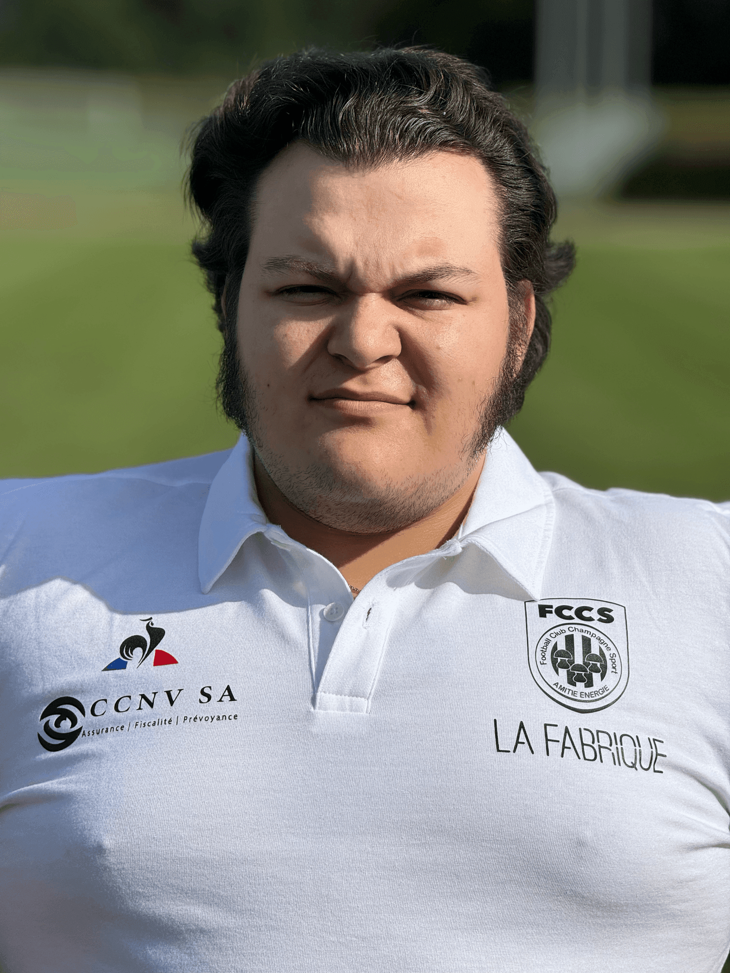 Person wearing a white polo shirt with multiple logos and text, standing outdoors in a sunny setting.