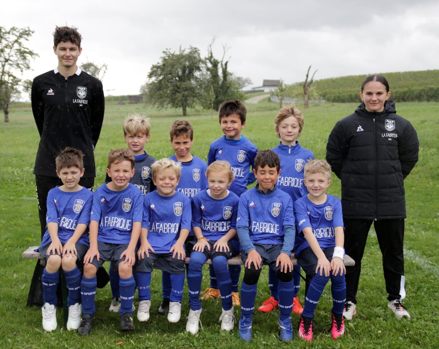 Youth soccer team posing outdoors with two coaches, wearing blue uniforms, on a green field.
