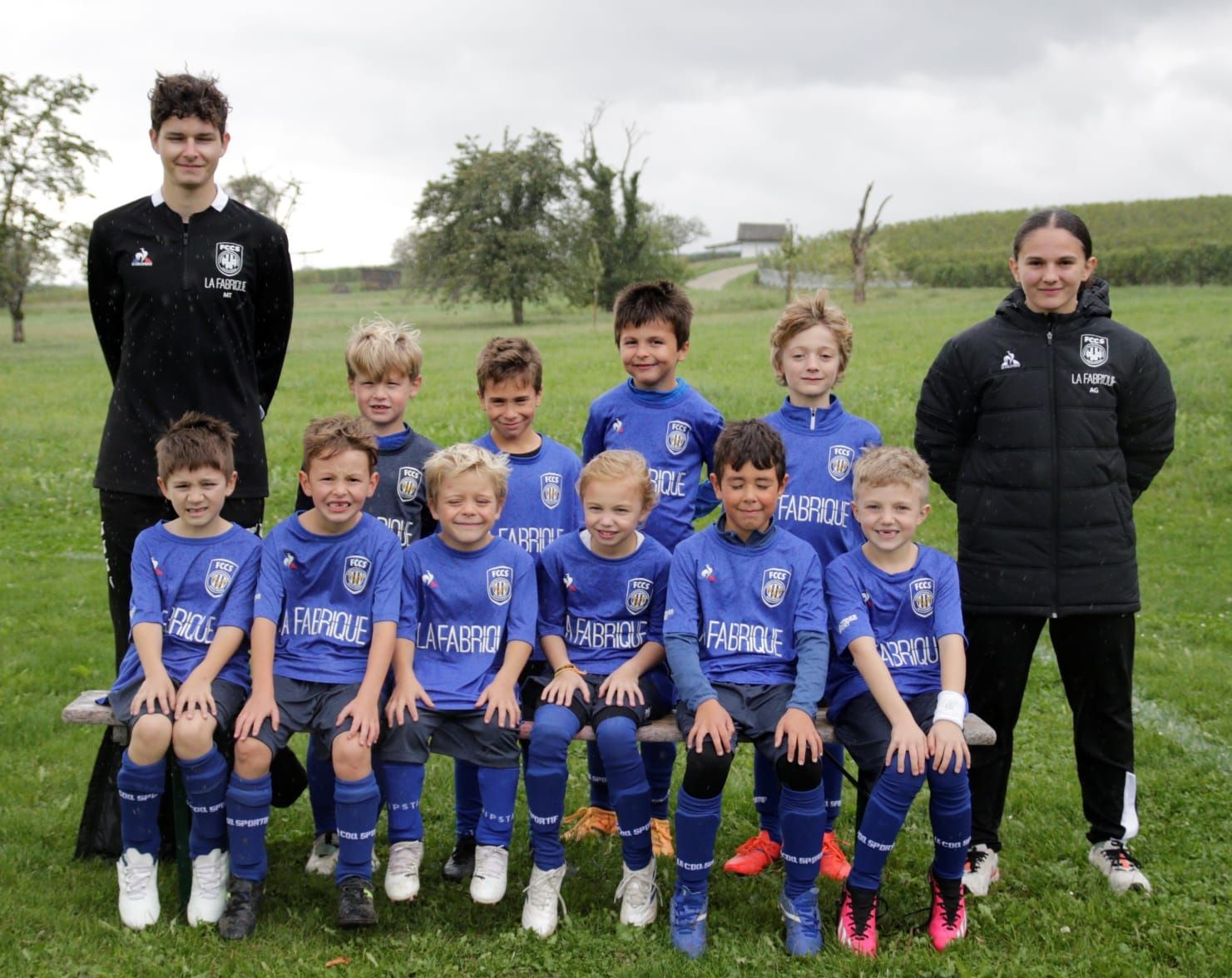 Youth soccer team posing outdoors with two coaches, wearing blue uniforms, on a green field.