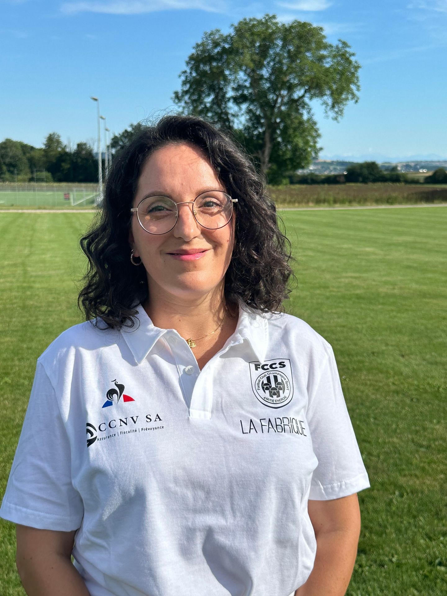 Person wearing a white polo shirt with logos standing on a grassy field under a clear blue sky.