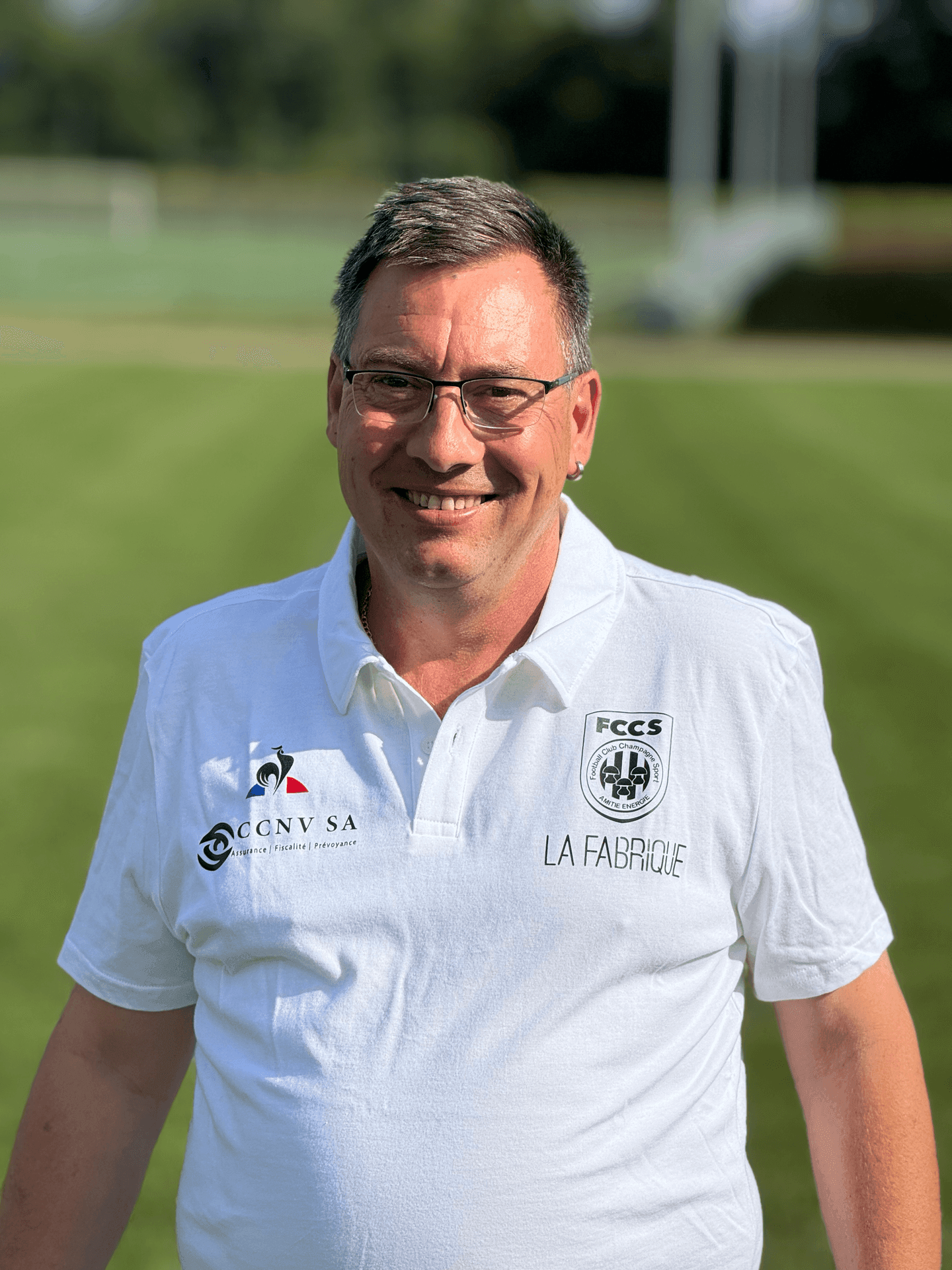 Man wearing glasses and a white polo shirt with logos, standing on a grassy field.