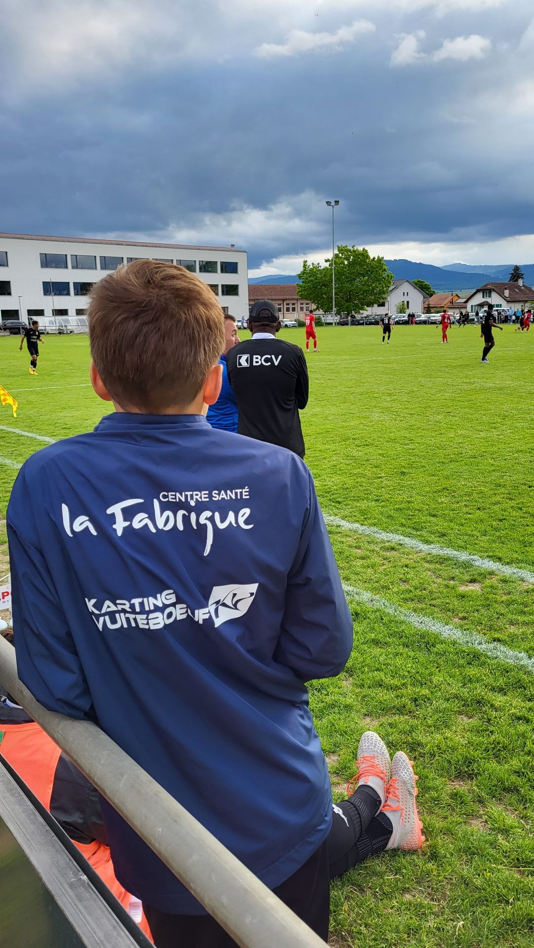 A person in a blue jacket watches a soccer game from the sidelines on a cloudy day.